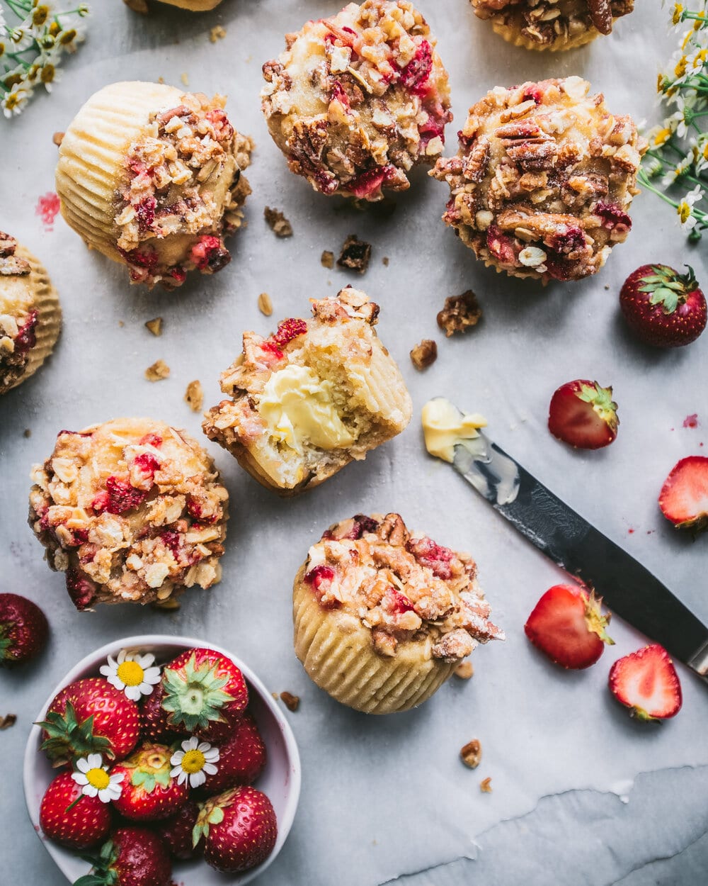 Overhead photo of finished muffins on a sheet of parchment paper next to strawberries, flowers and a knife.