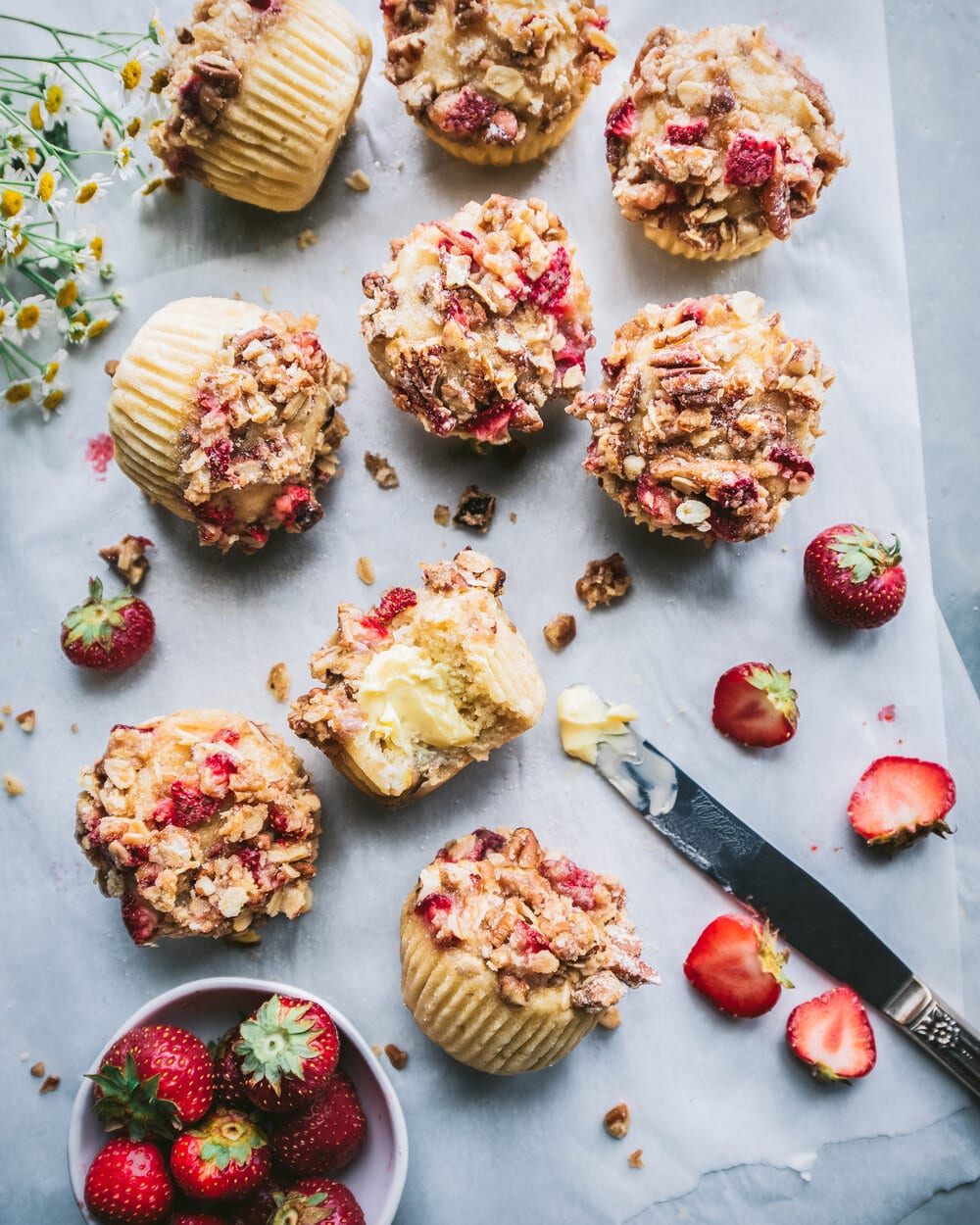 Overhead photo of finished muffins on a sheet of parchment paper next to flowers, strawberries and a knife.