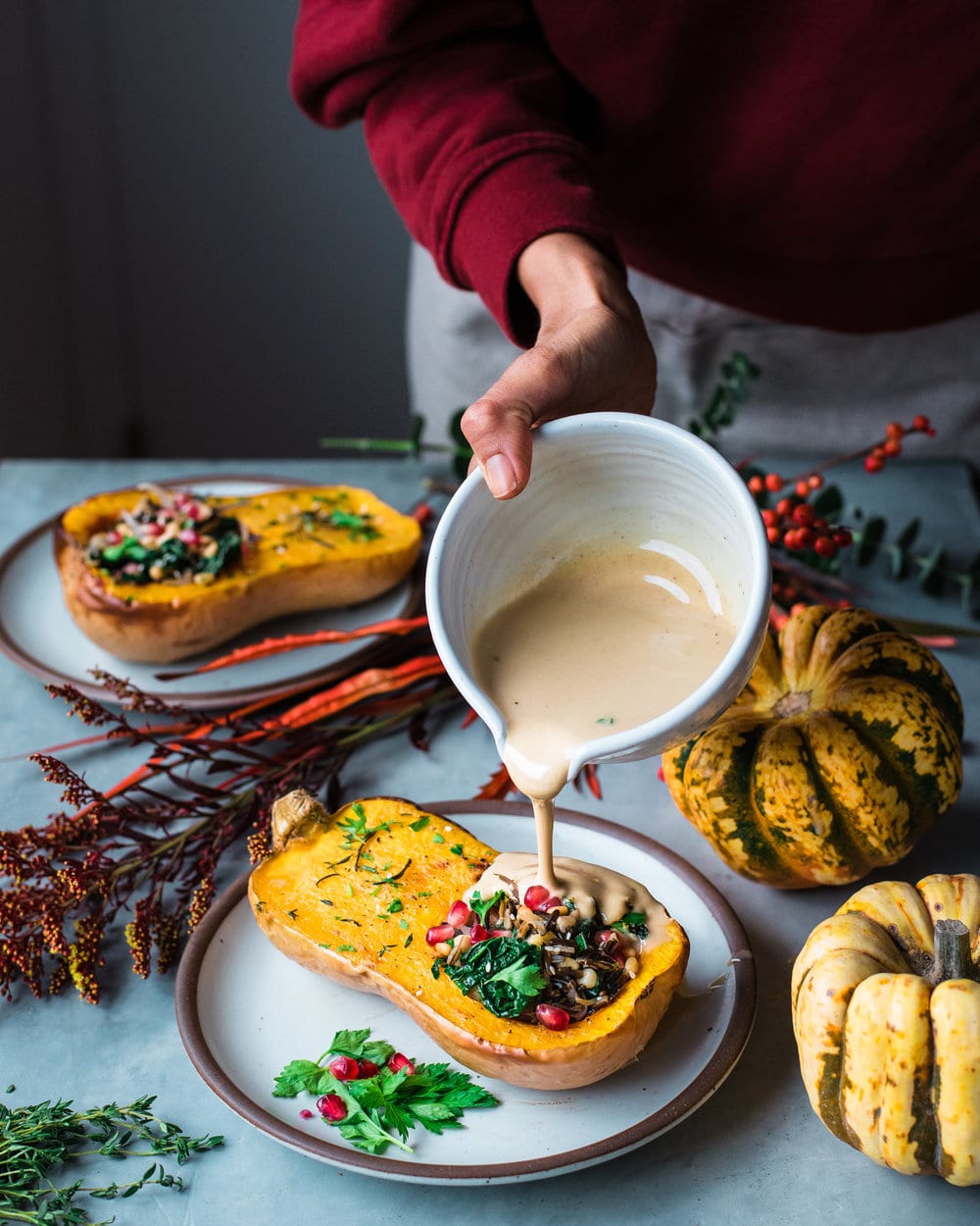 Woman pouring tahini sauce onto half a squash on a grey fall-decorated table.