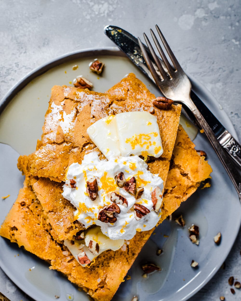 Stack of three sheet pancakes with butter, whipped cream and orange zest on a plate with utensils.