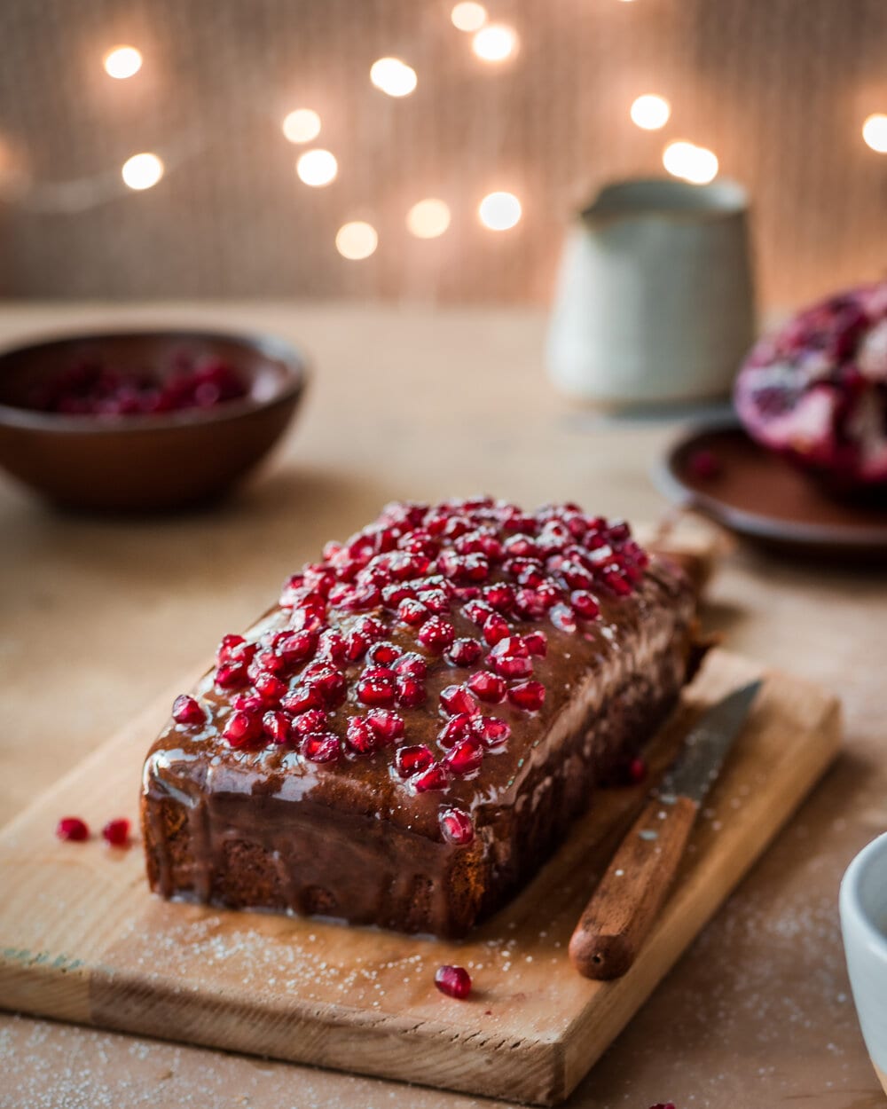Gingerbread cake with icing and pomegranate seeds on a wood cutting board.