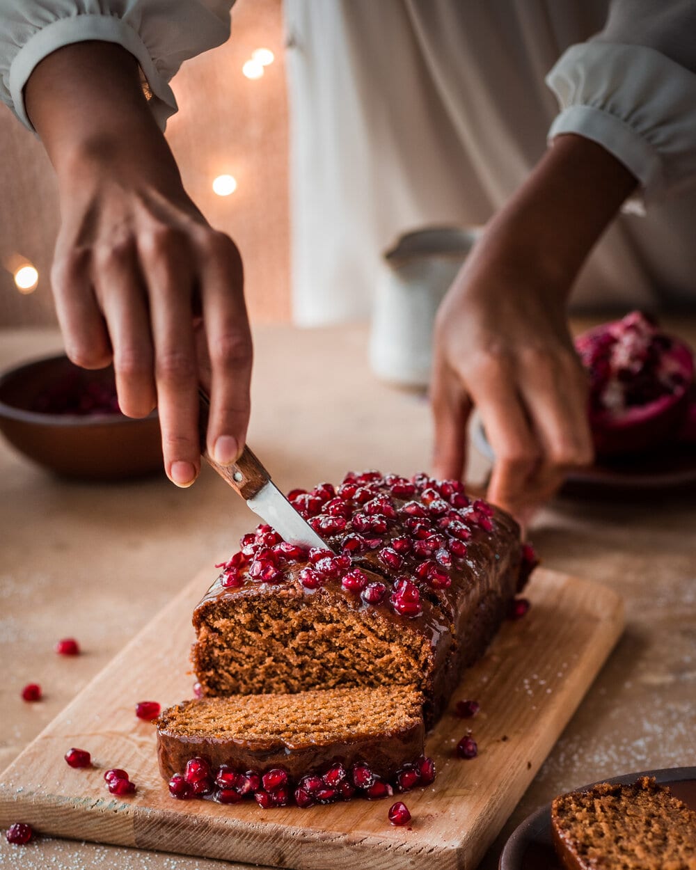 Woman slicing gingerbread cake with pomegranate seeds on a wood cutting board.