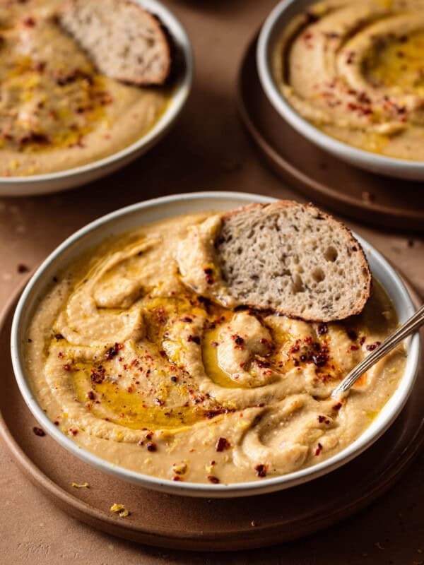 bowl of creamy vegan cauliflower soup with spoon dug in and bread dipped in on a brown table with two bowls in back.