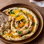 a piece of bread dunked into a creamy hummus swirled onto a plate with olive oil, paprika, parsley