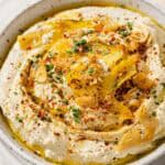 side view of a bowl of hummus with lemon garlic topping next to pita triangles.