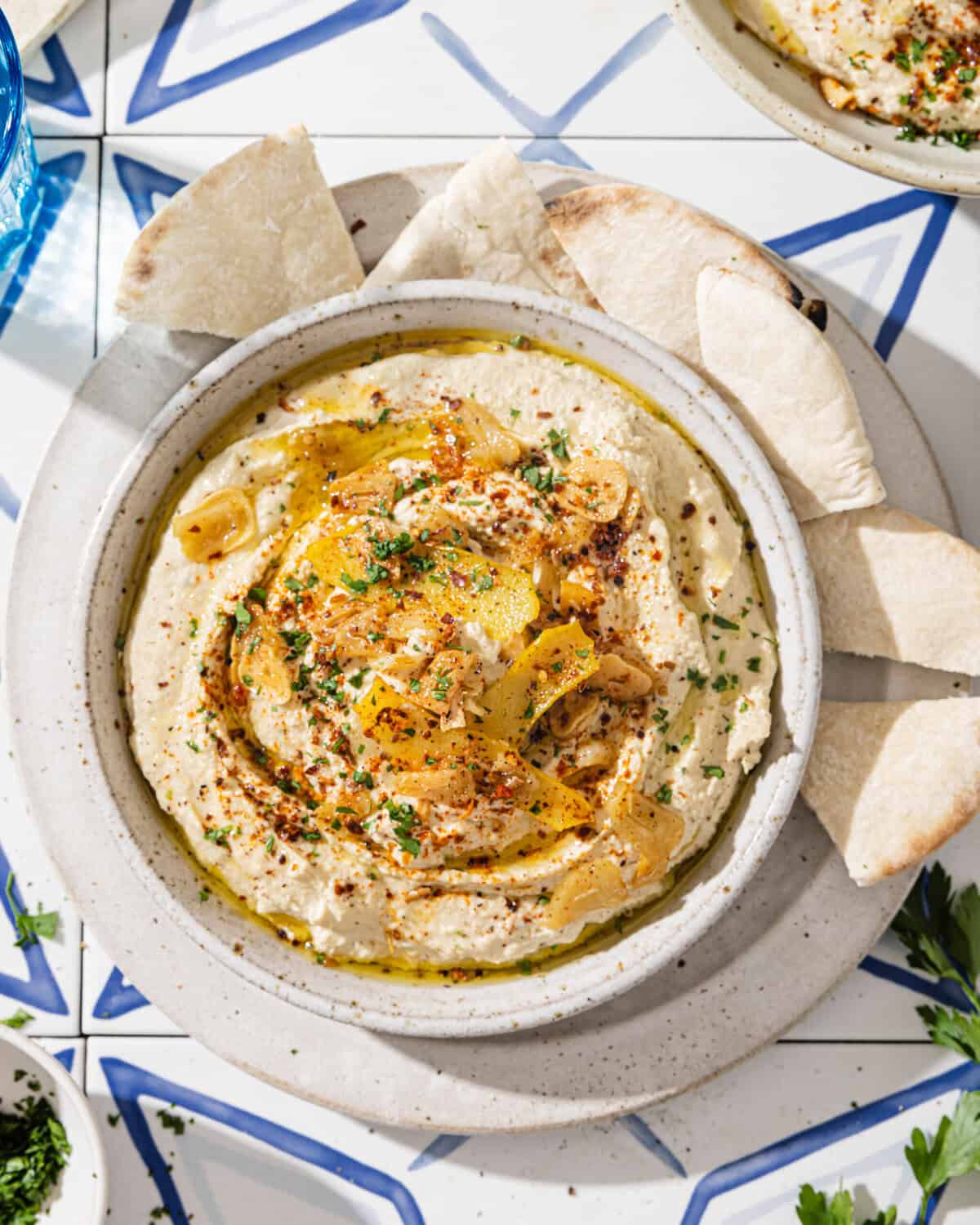 overhead view of hummus with lemon garlic topping in a bowl next to pita on white & blue tiled surface.