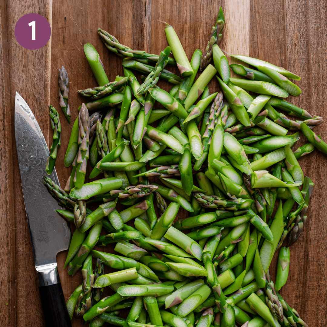 thinly sliced asparagus pieces cut on the diagonal on a wooden cutting board with knife.