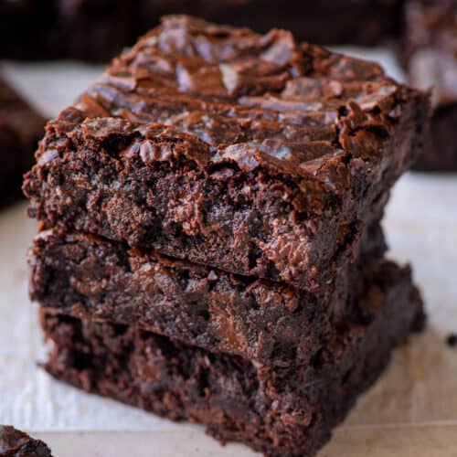 stack of three fudgy, chewy vegan brownies on parchment paper with more brownies in the background.