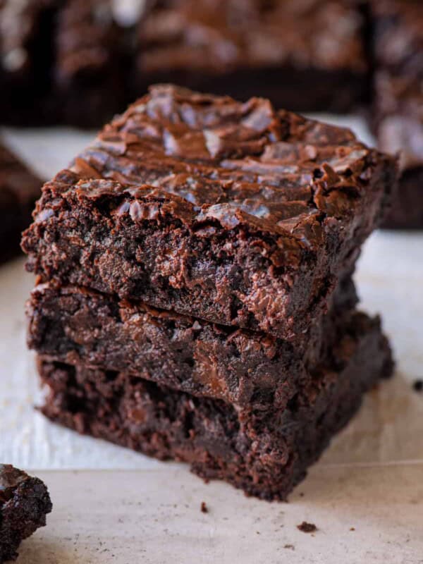 stack of three fudgy, chewy vegan brownies on parchment paper with more brownies in the background.