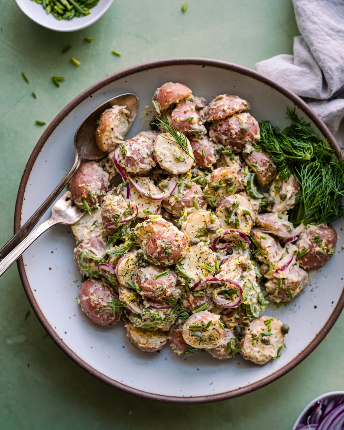 creamy vegan potato salad with dilll in a white bowl with garnishes on a green backdrop