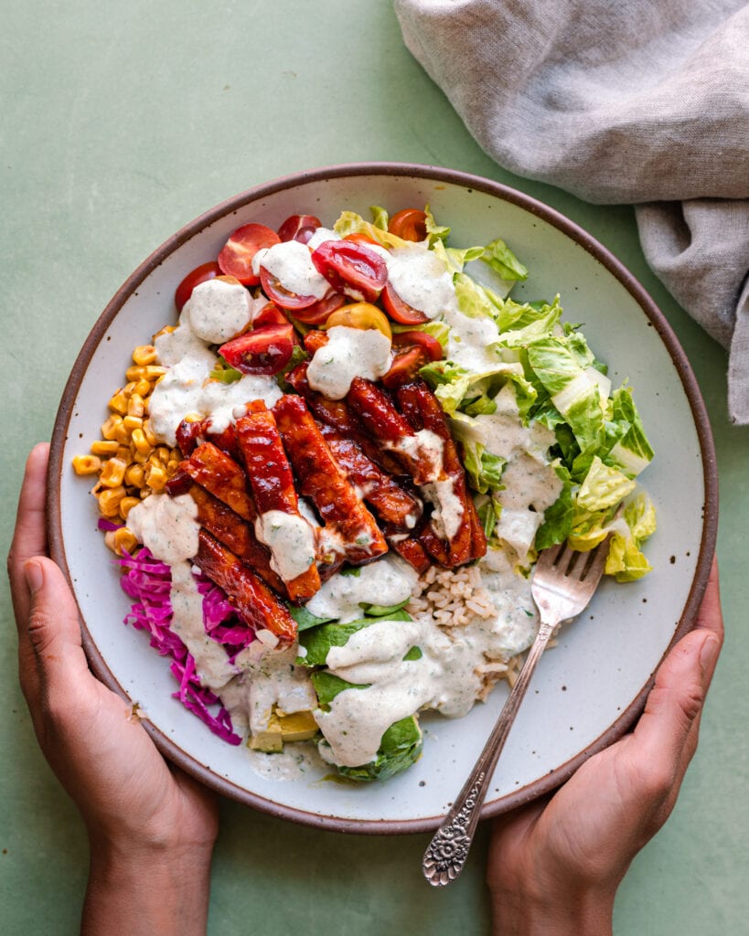 woman's hands holding white bowl of bbq tempeh bowl