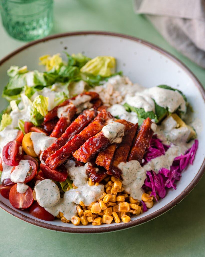 BBQ tempeh bowl with ranch dressing, lettuce, corn and avocado on green surface