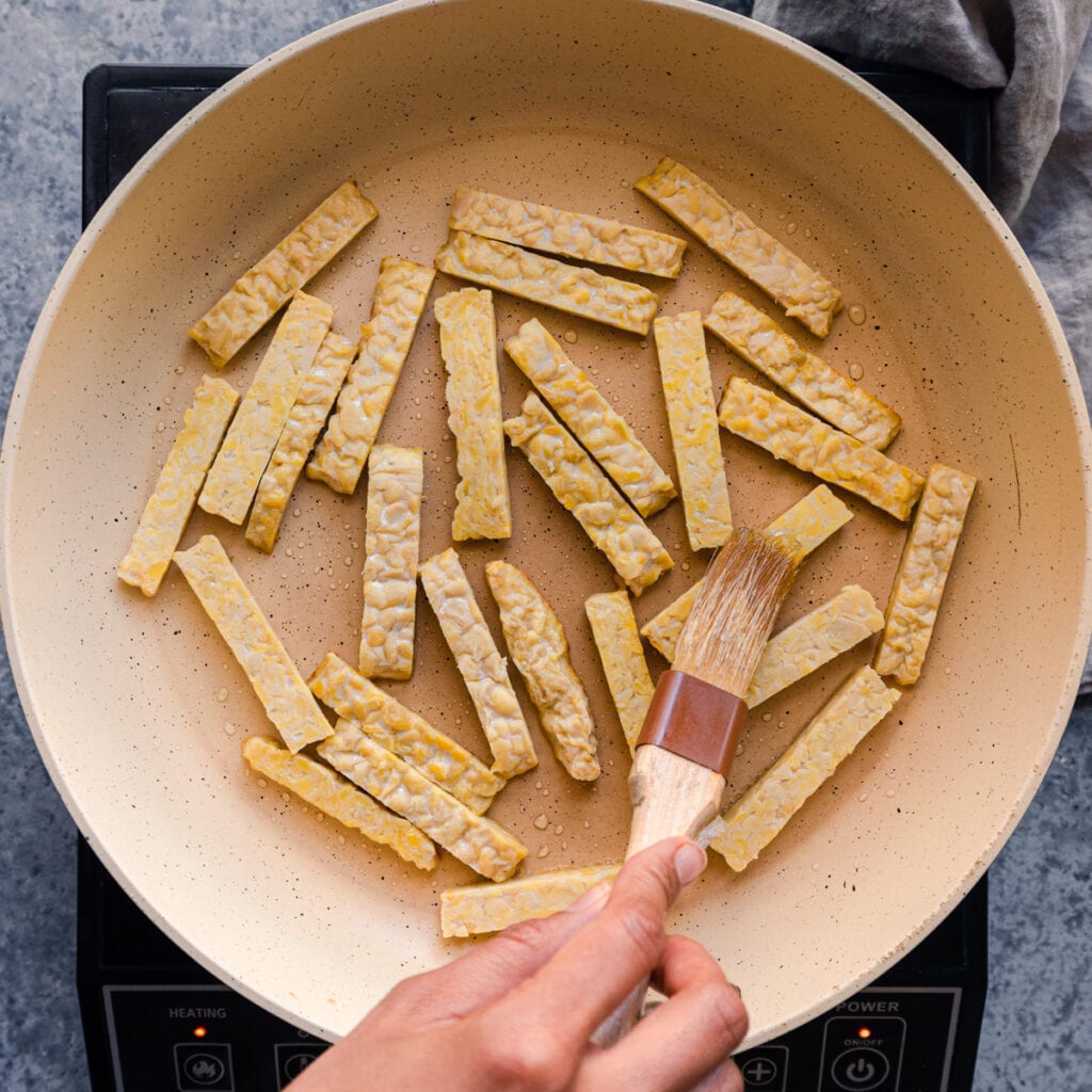 brushing oil on tempeh in frying pan