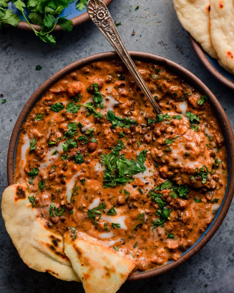 bowl of creamy vegan dal makhani with spoon and naan on blue background