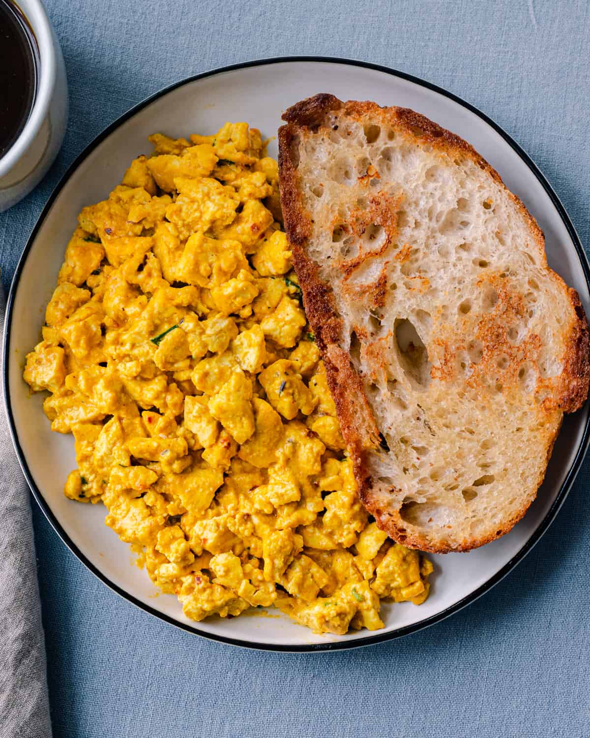 creamy tofu scramble piled high on a white plate with a large slice of toasted bread next to it on a blue tablecloth. 
