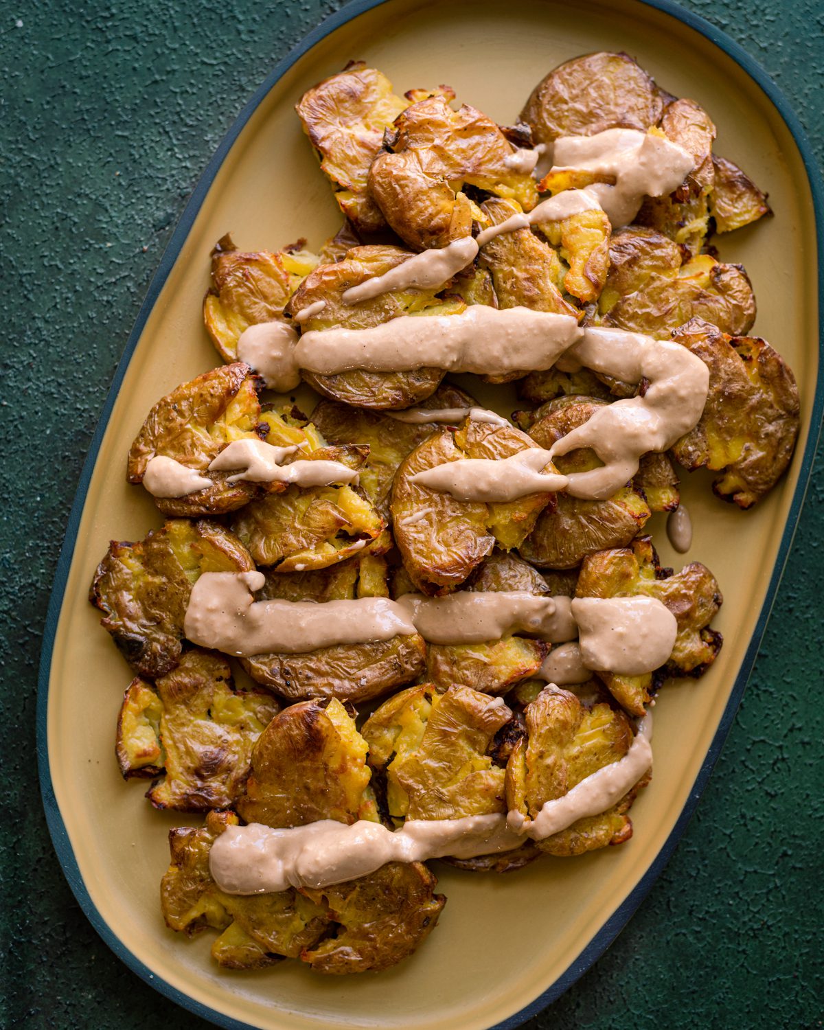 crispy smashed potatoes with ginger-tahini sauce drizzled on top on a yellow serving platter