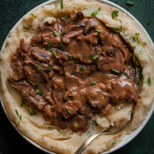 a big pile of vegan gravy with mushrooms on a bowl of mashed potatoes with a spoon dug in on a green table.
