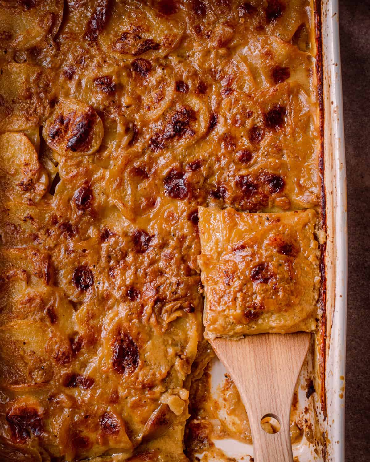 cheesy vegan scalloped potatoes in casserole pan with slice being lifted up by a spatula