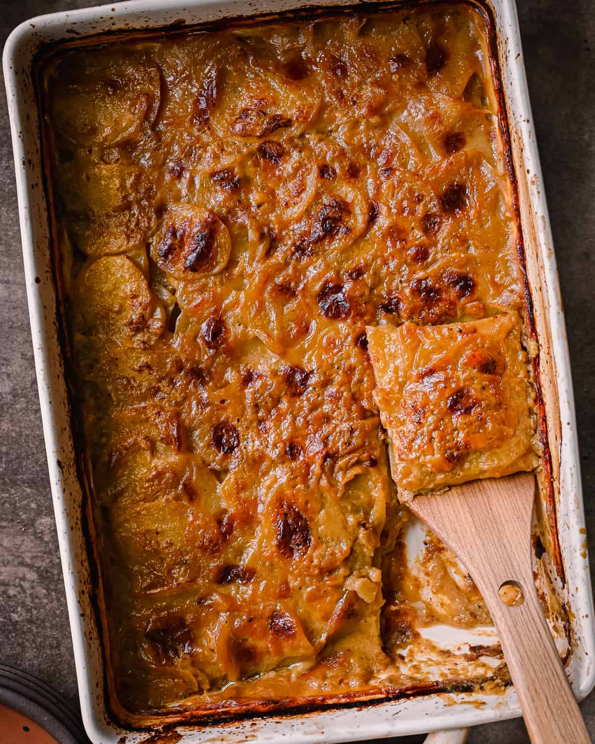 cheesy vegan scalloped potatoes in casserole pan with slice being lifted up by a spatula