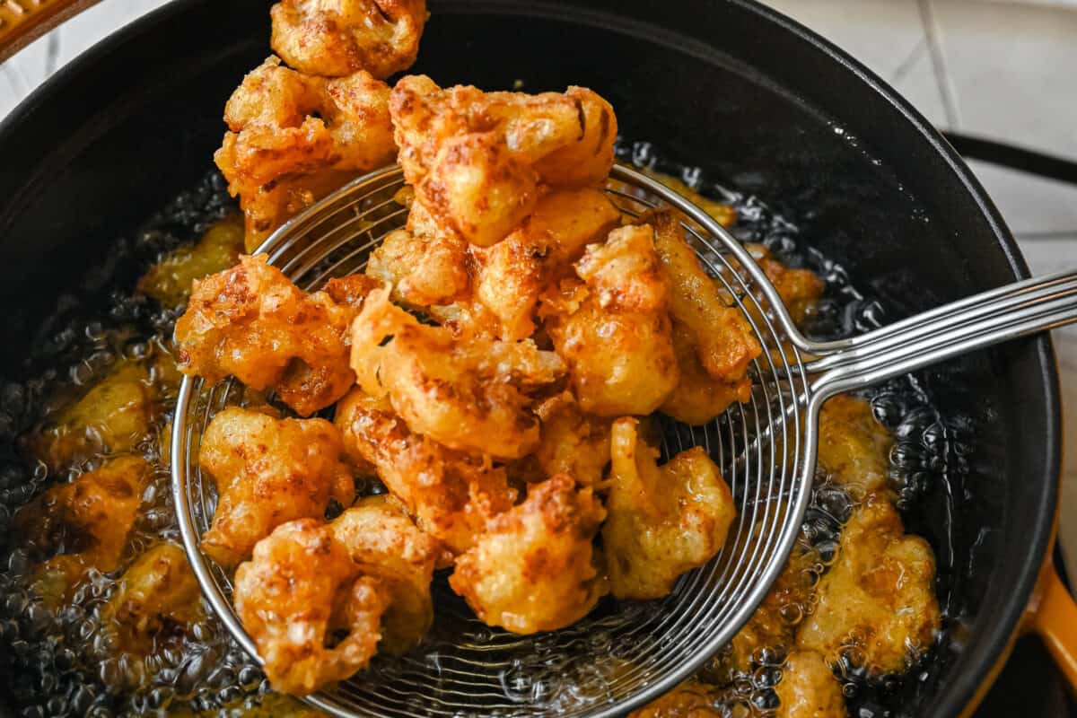 scooping fried cauliflower florets in a spider tool above a pan of hot oil