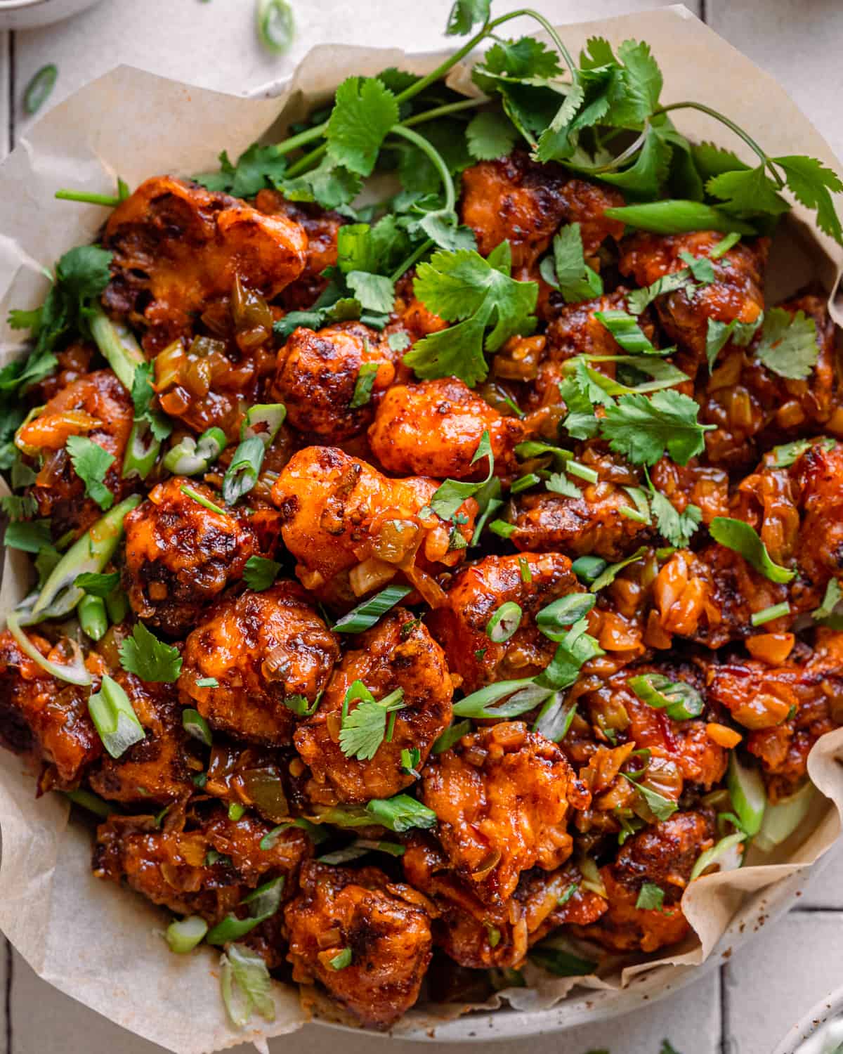 Gobi Manchurian in a parchment paper lined bowl on a white tiled surface, garnished with cilantro and scallions