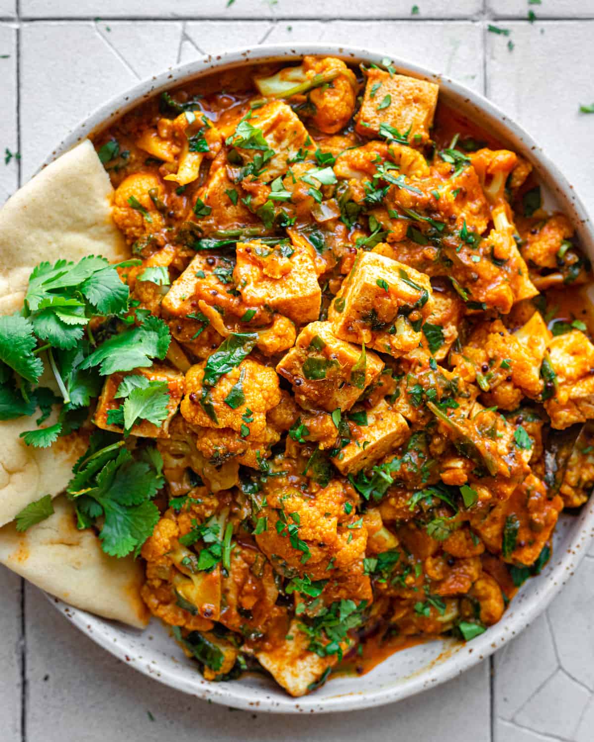 vegan curry with tofu in a white plate with naan and cilantro on white tiled surface.
