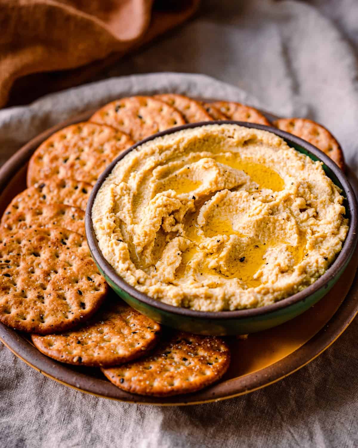 bowl of vegan ricotta with olive oil drizzled on top on a plate with crackers, on a linen towel