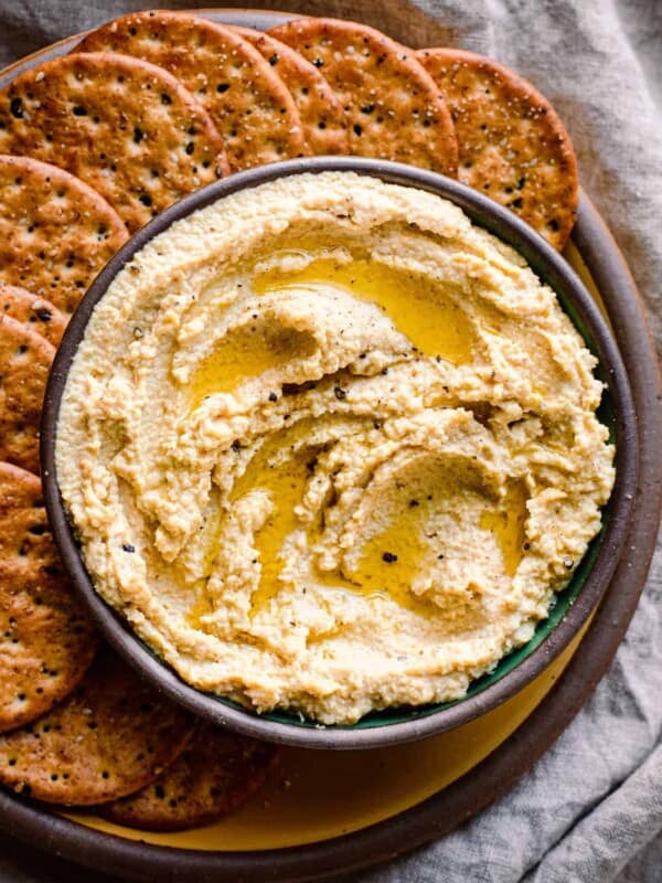 bowl of vegan ricotta with olive oil drizzled on top on a plate with crackers, on a linen towel.