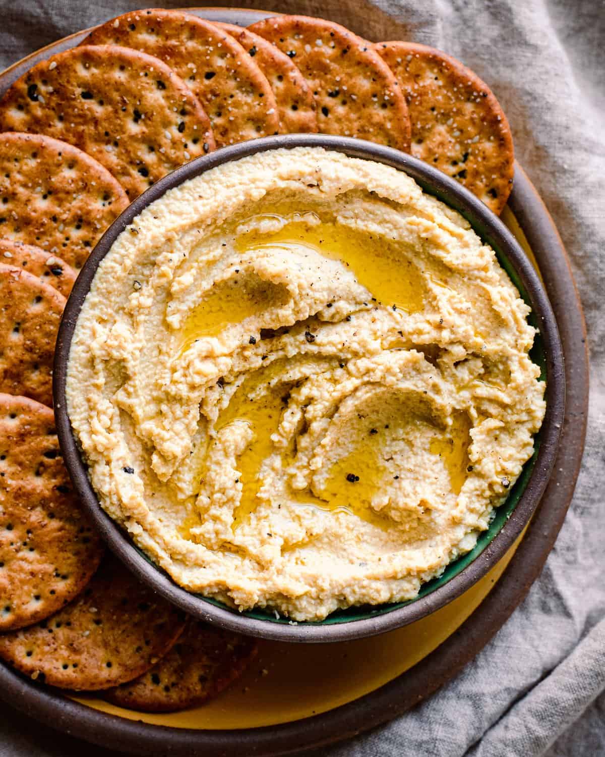 bowl of vegan ricotta with olive oil drizzled on top on a plate with crackers, on a linen towel.