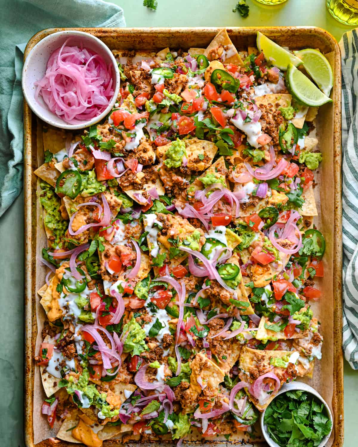 vegan nachos on a tray with pickled onions, lime wedges, and cilantro, on a green backdrop with glasses of beer