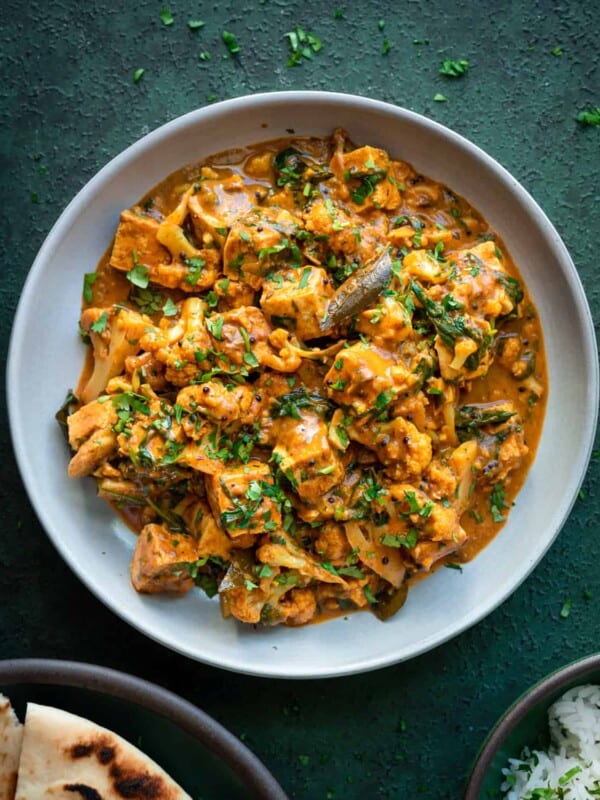 vegan curry with tofu in a white plate with naan and cilantro on a dark green surface.