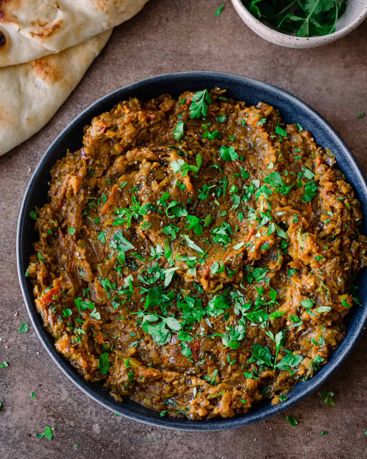 overhead shot of baingan bharta in a shallow navy bowl with naan on the side, garnished with cilantro