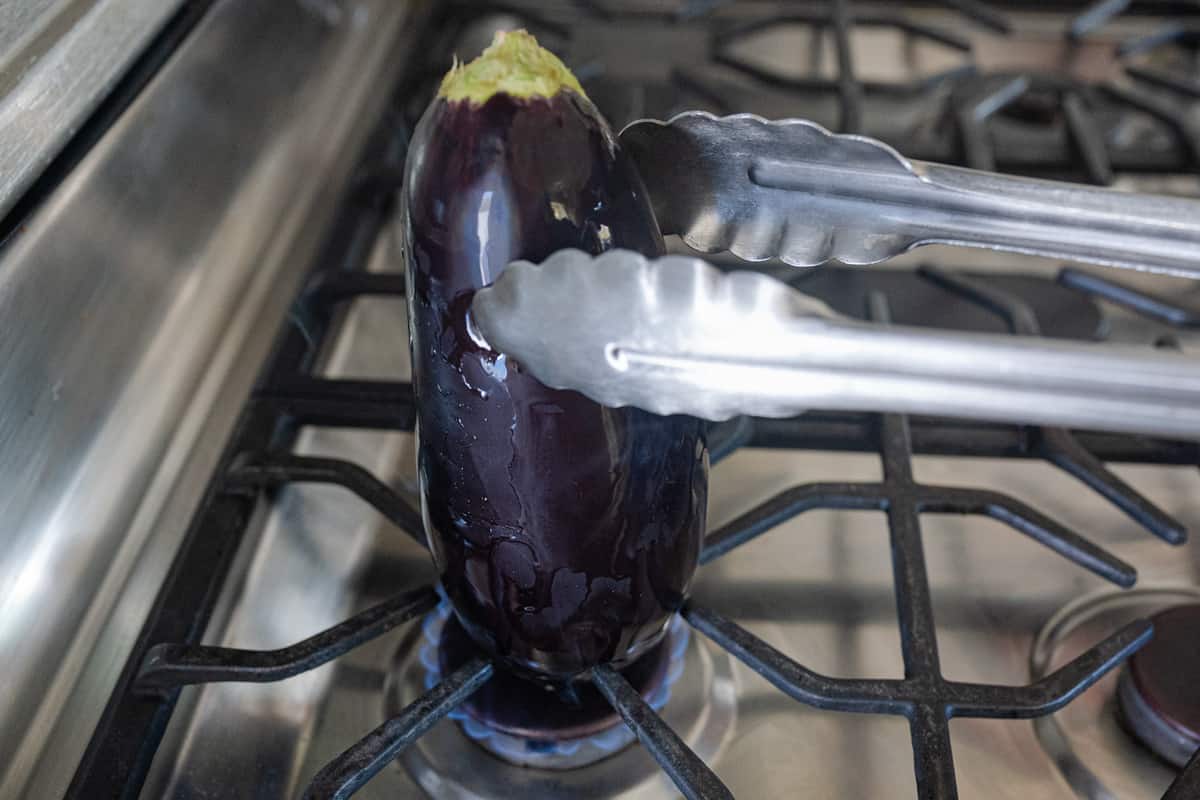 tongs holding whole eggplant roasting on open gas flame for baingan bharta