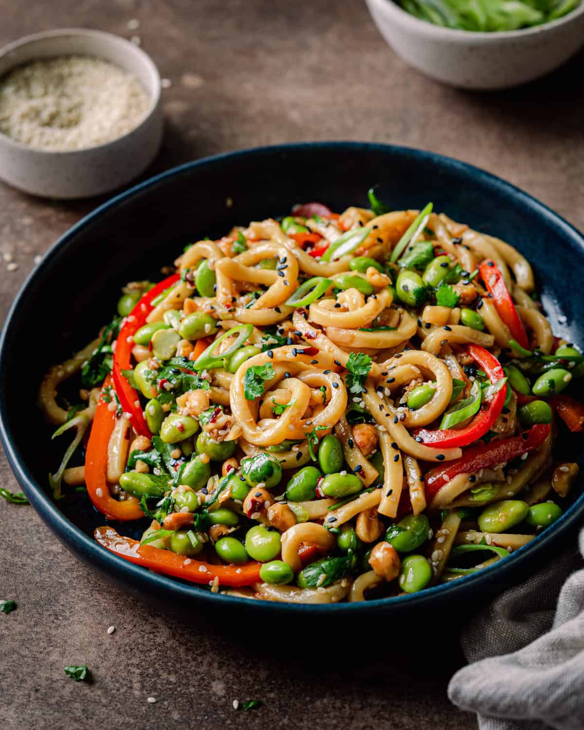 vegan noodles with chili garlic sauce and bell peppers in navy blue bowl on brown backdrop, with bowl of scallions and bowl of sesame seeds