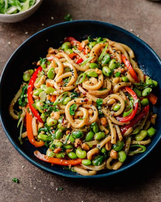 vegan noodles with chili garlic sauce and bell peppers in navy blue bowl on brown backdrop, with bowl of sesame seeds