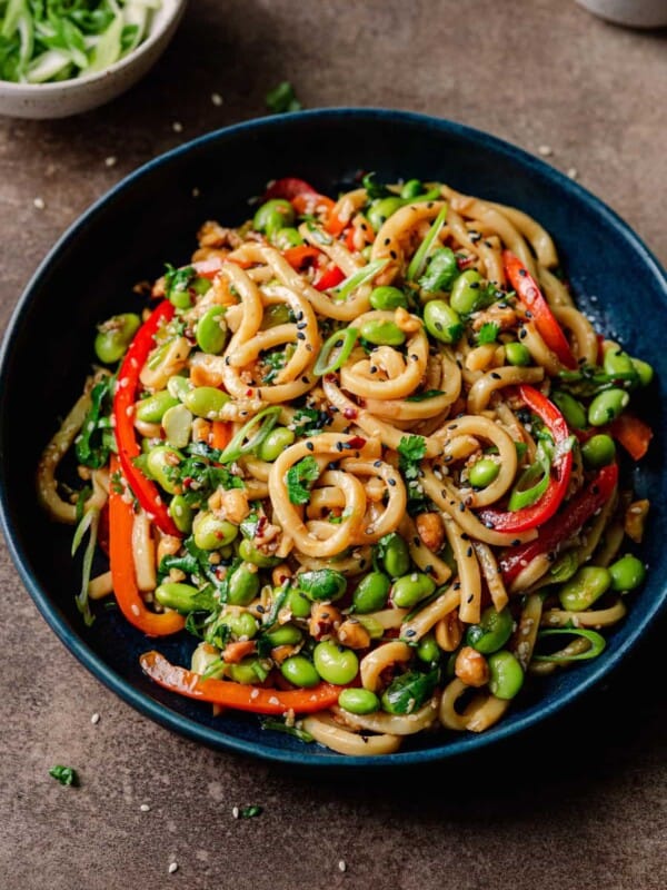 vegan noodles with chili garlic sauce and bell peppers in navy blue bowl on brown backdrop, with bowl of sesame seeds.