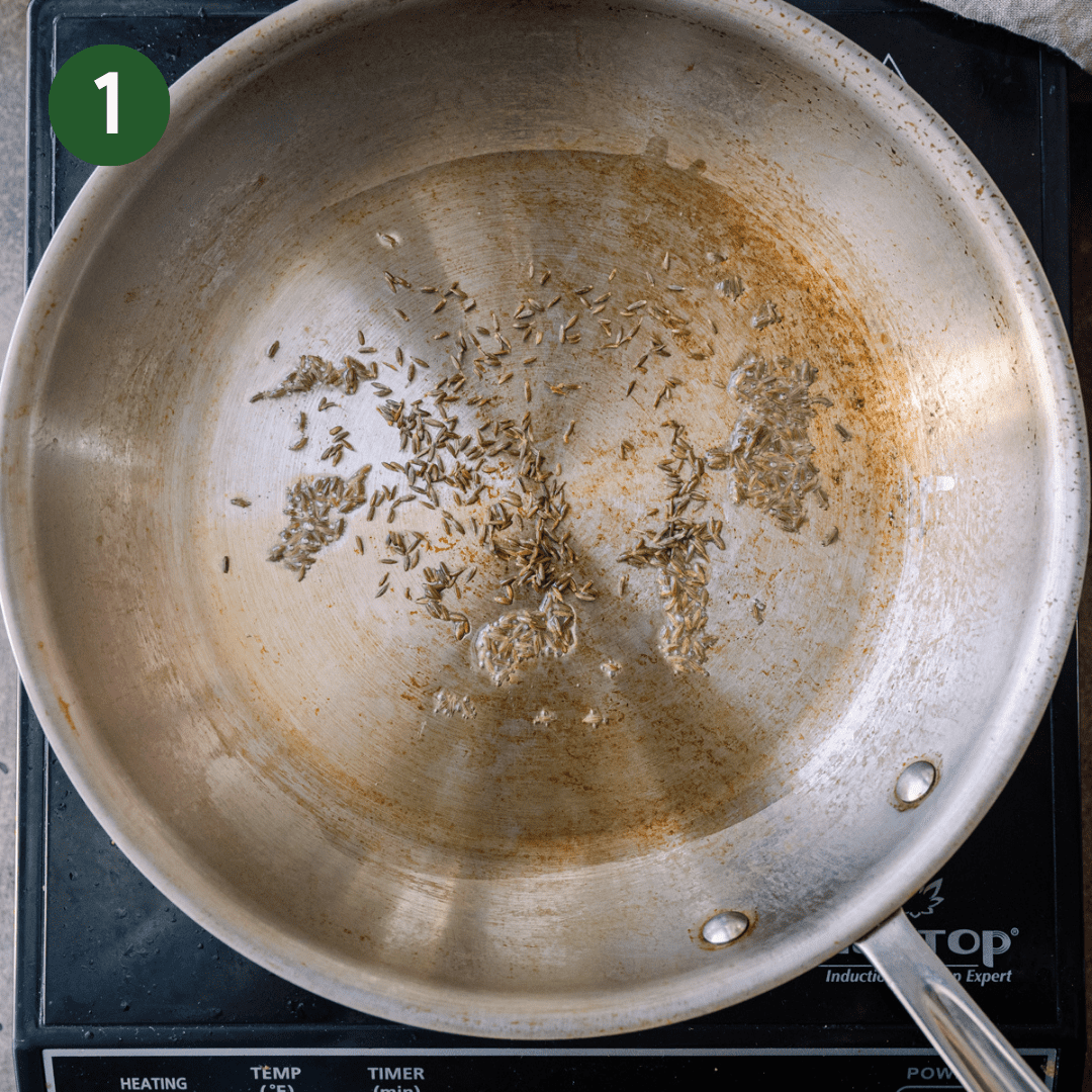 cumin seeds frying in oil in a stainless steel skillet.