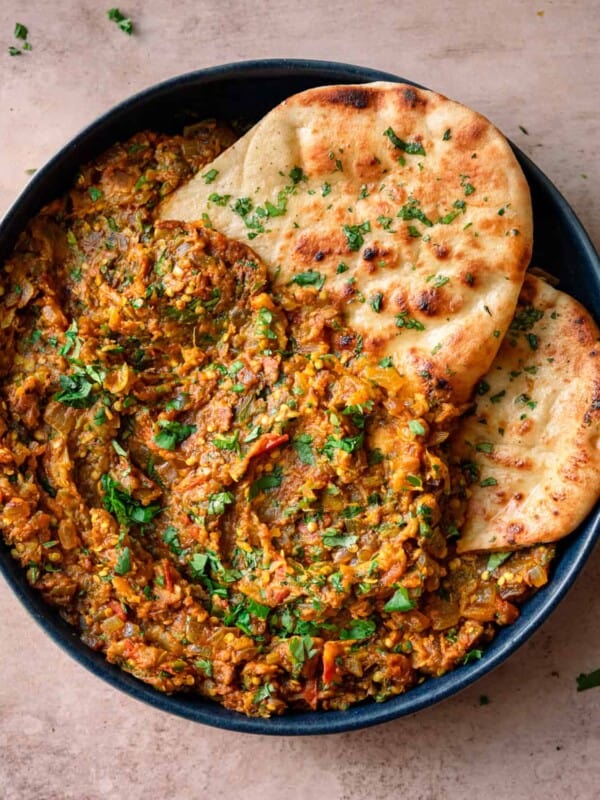 baingan bharta in a navy blue dish with cilantro naan tucked in on a light pink table.