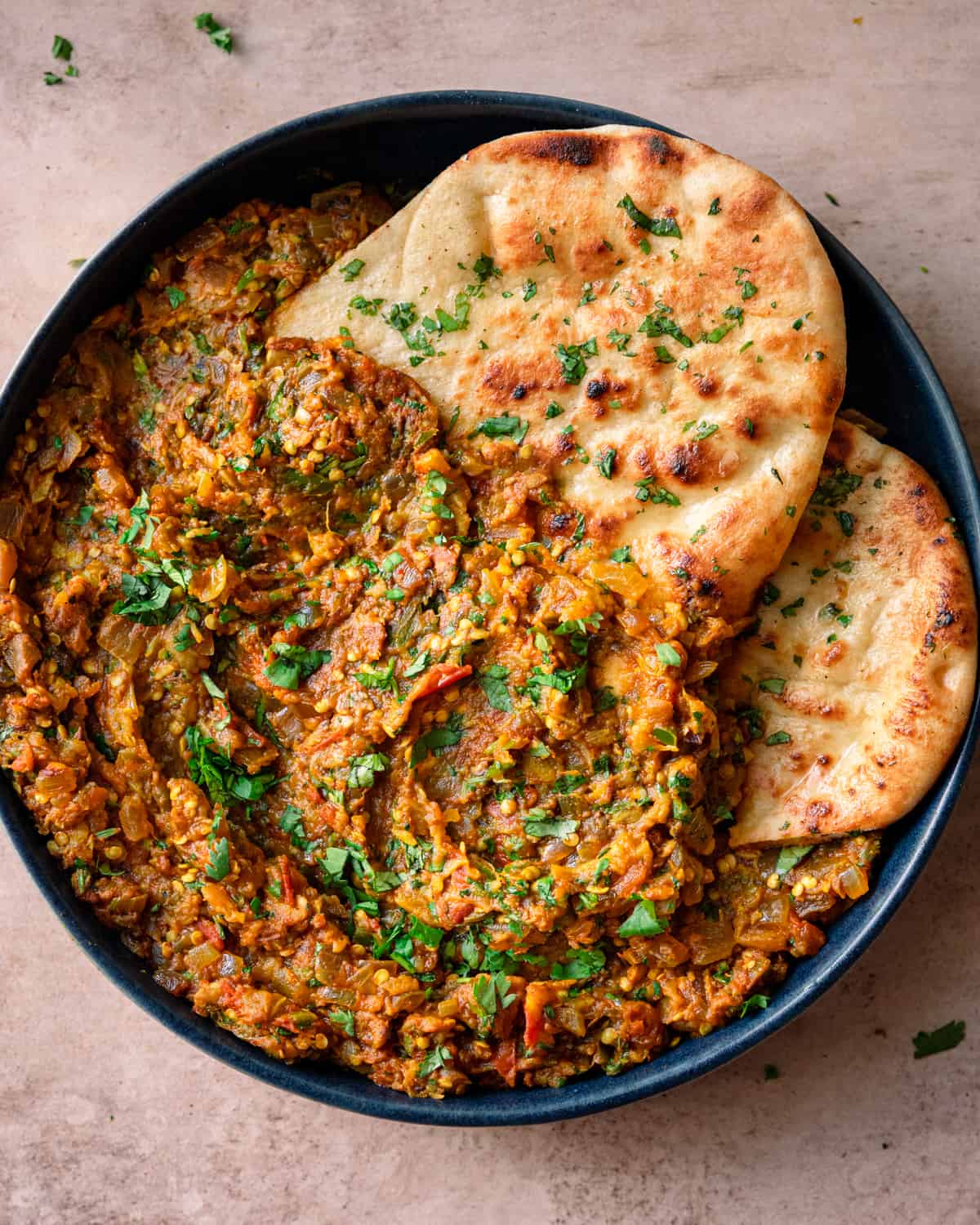 baingan bharta in a navy blue dish with cilantro naan tucked in on a light pink table.