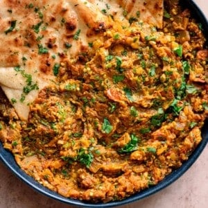 baingan bharta in a navy blue dish with cilantro naan tucked in on a light pink table.