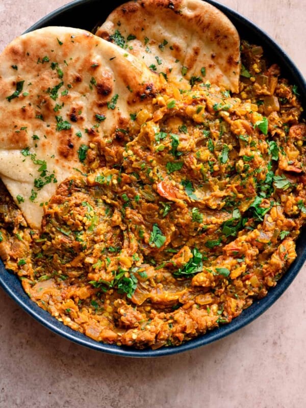baingan bharta in a navy blue dish with cilantro naan tucked in on a light pink table.