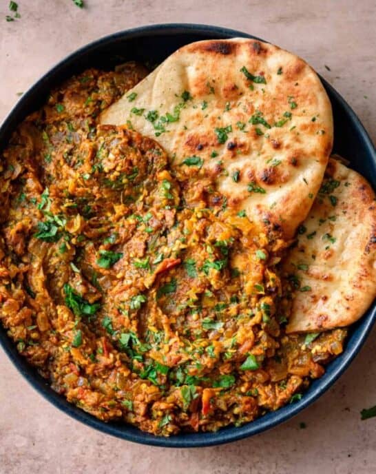 baingan bharta in a navy blue dish with cilantro naan tucked in on a light pink table.