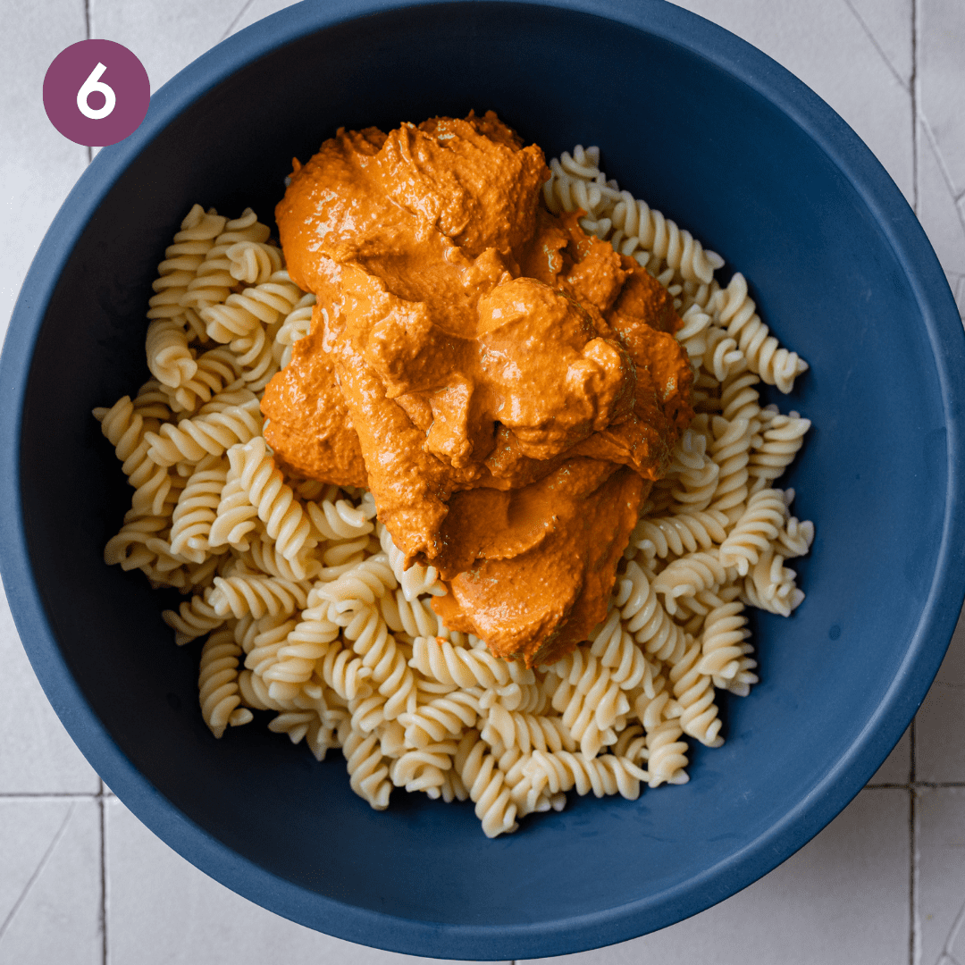 big scoop of roasted red pepper sauce on top of cooked pasta in a blue bowl.