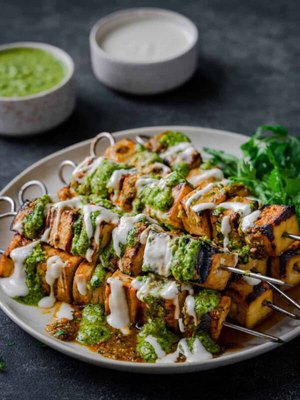 Grilled Tofu Skewers on a plate, covered in creamy white sauce, pesto, and with cilantro.