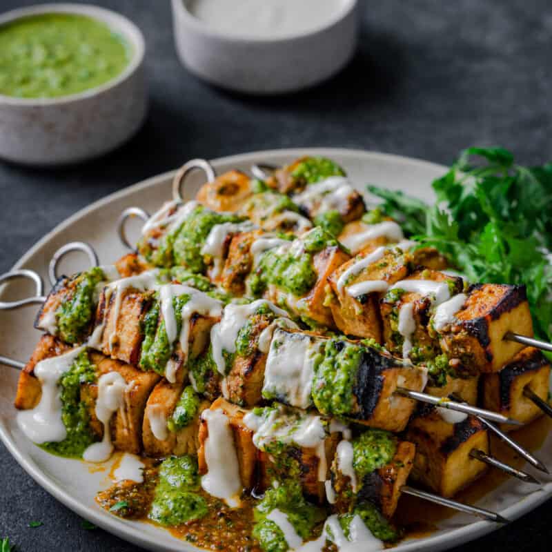 Grilled Tofu Skewers on a plate, covered in creamy white sauce, pesto, and with cilantro.