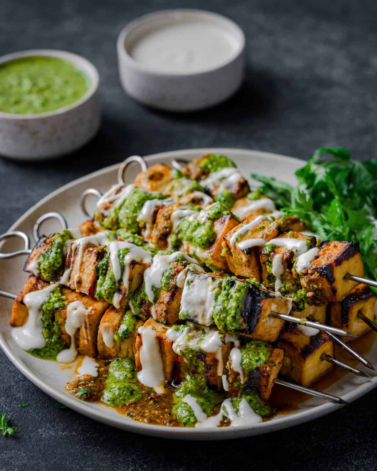 Grilled Tofu Skewers on a plate, covered in creamy white sauce, pesto, and with cilantro.