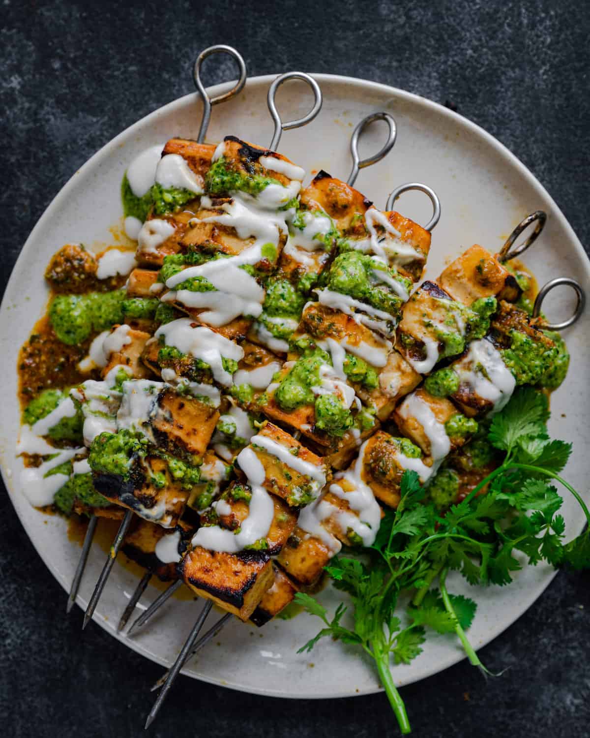 Grilled Tofu Skewers on a plate, covered in creamy white sauce, pesto, and with cilantro on a dark backdrop.