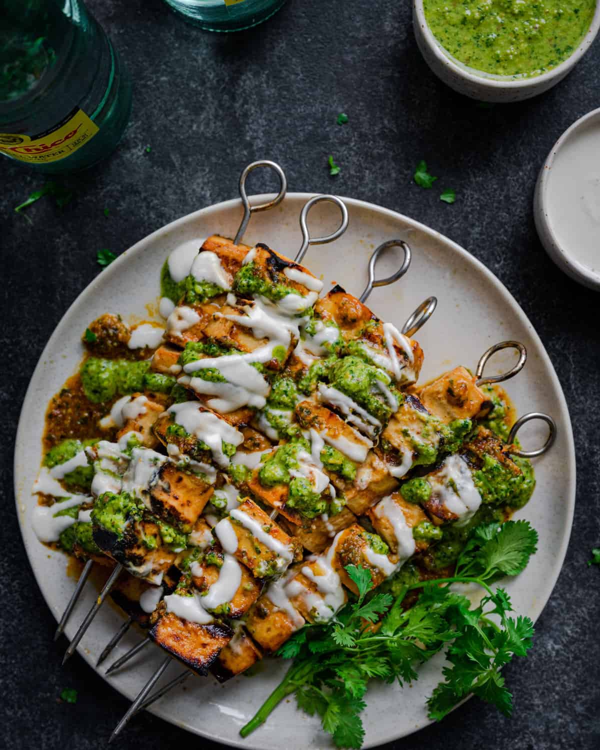 Grilled Tofu Skewers on a plate, covered in creamy white sauce, pesto, and with cilantro on a dark backdrop.