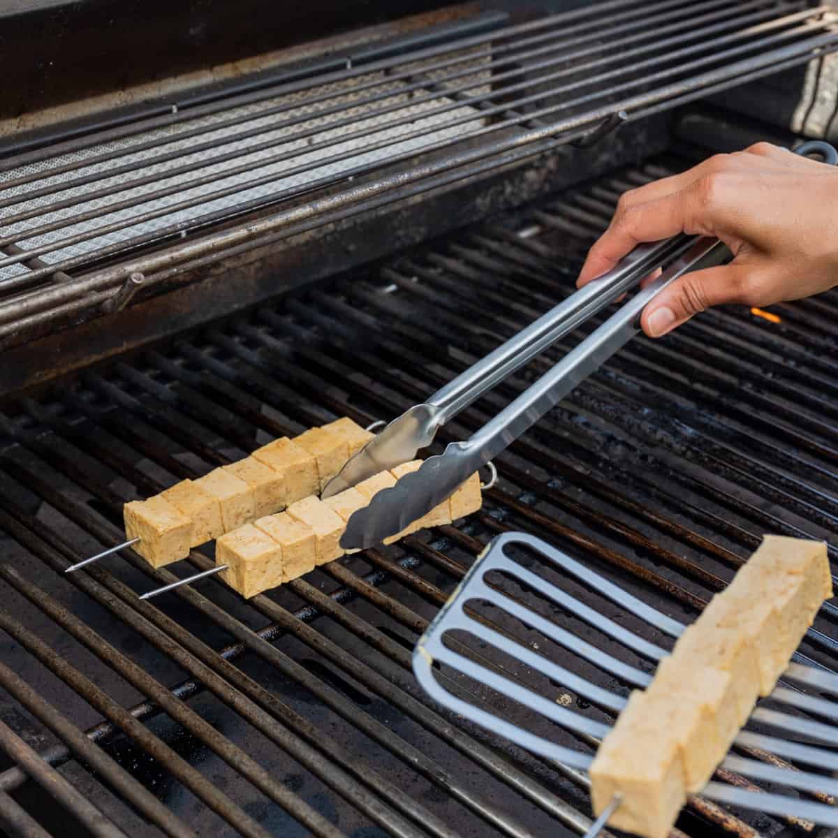 Woman's hands arranging marinated tofu skewers onto an outdoor gas grill.