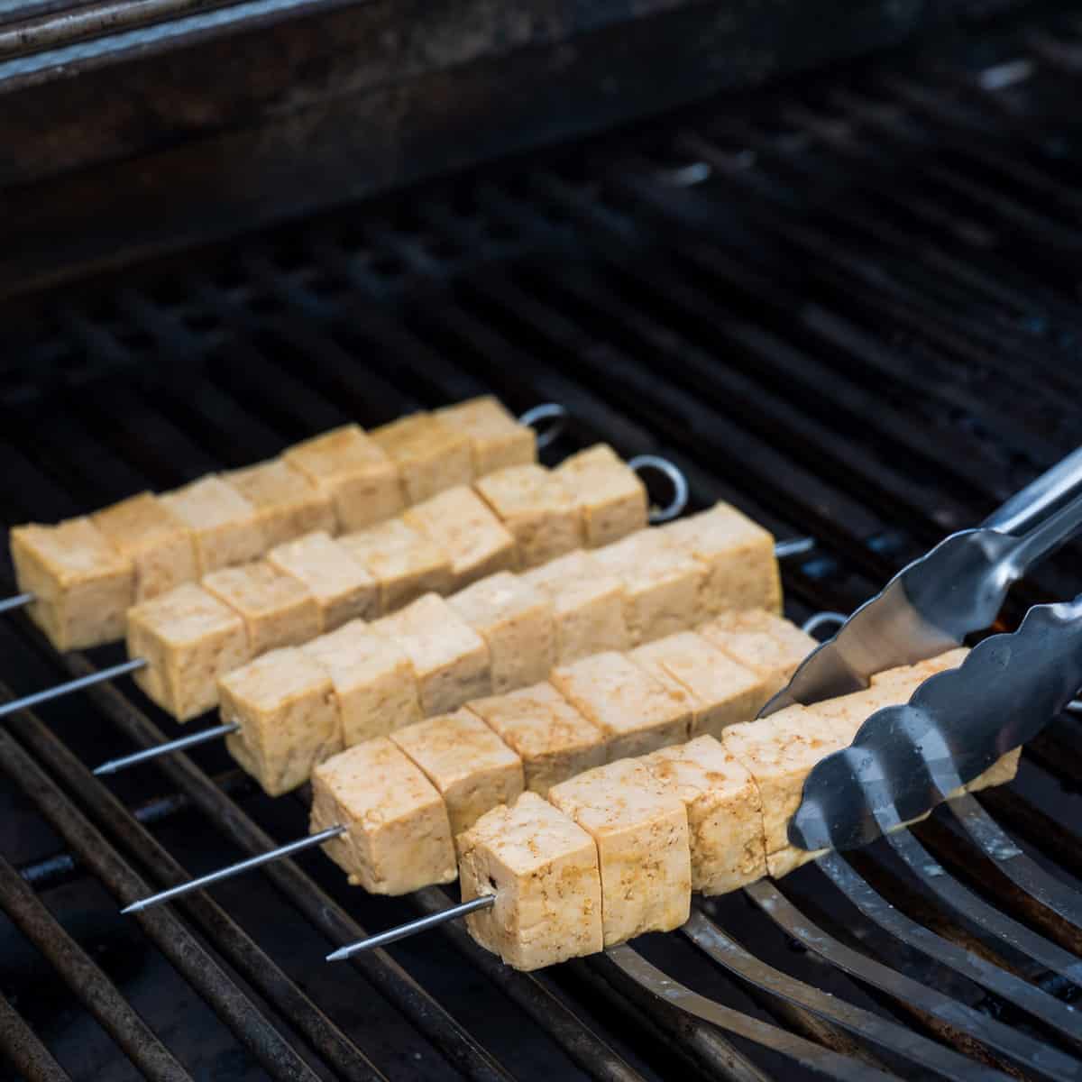 Tongs arranging tofu skewers on an outdoor gas grill.